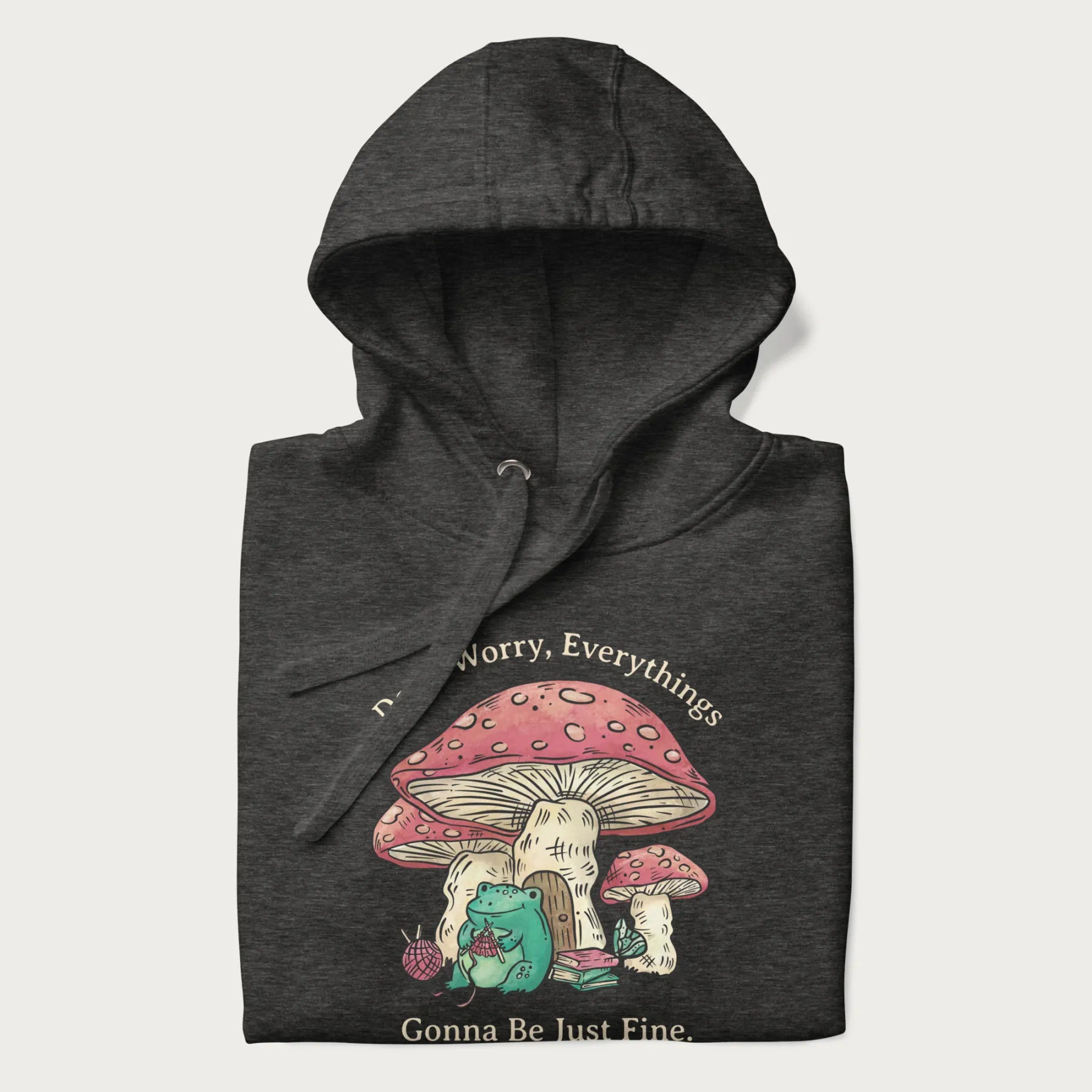 Folded dark grey hoodie with a graphic of a frog knitting under a large mushroom with the text 'Don't Worry, Everything's Gonna Be Just Fine'.