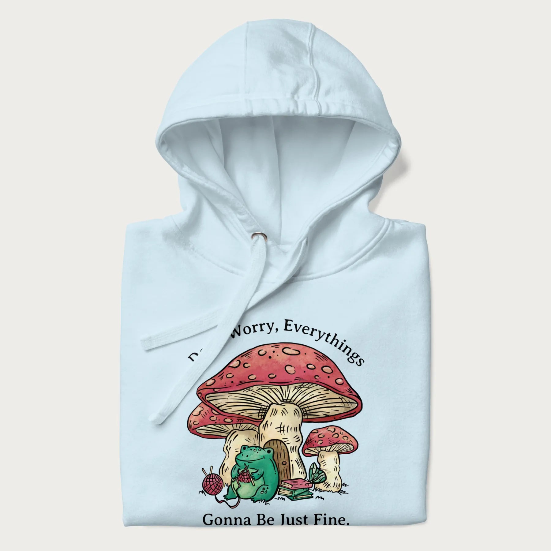 Folded light blue hoodie with a graphic of a frog knitting under a large mushroom with the text 'Don't Worry, Everything's Gonna Be Just Fine'.