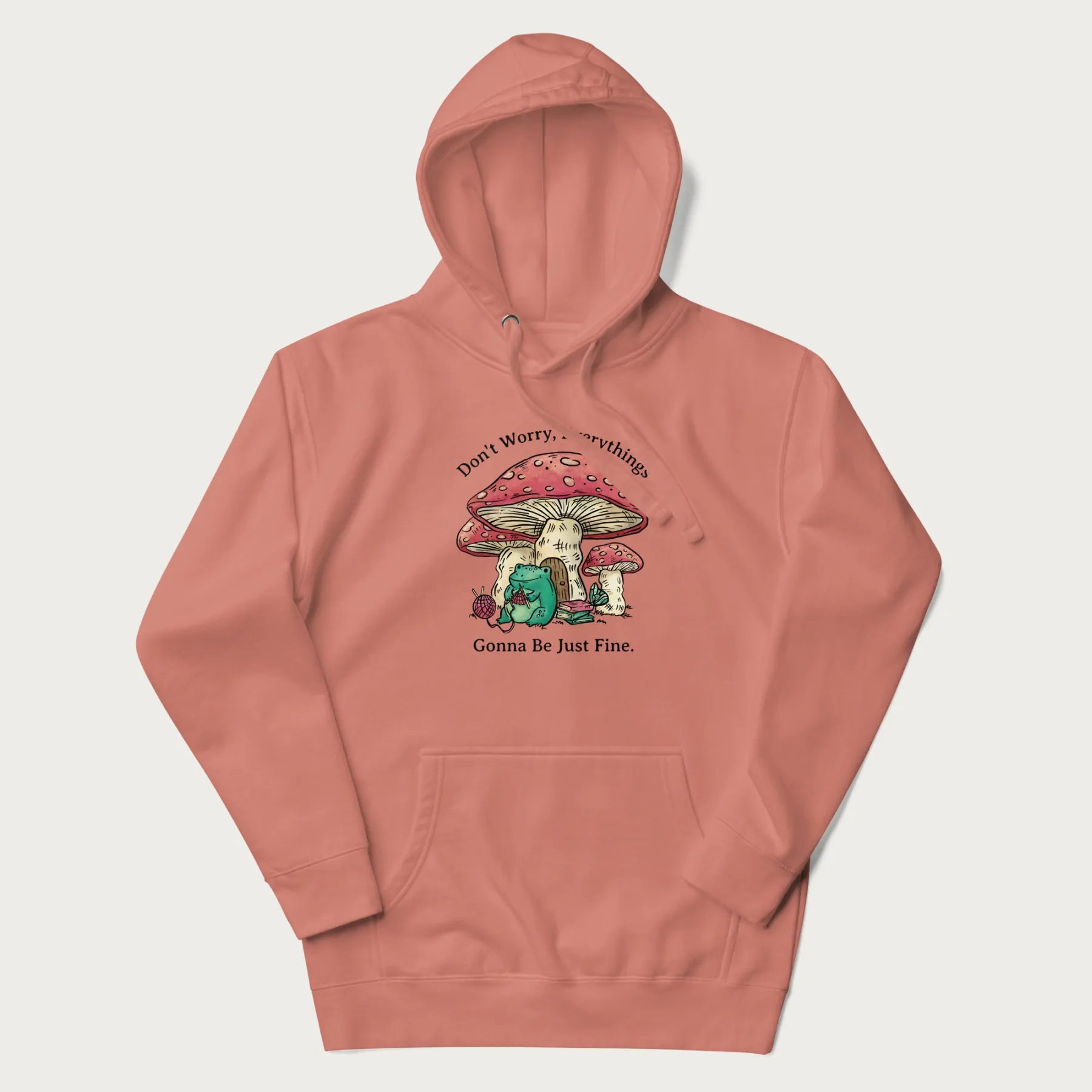 Light pink hoodie with a graphic of a frog knitting under a large mushroom with the text 'Don't Worry, Everything's Gonna Be Just Fine'.