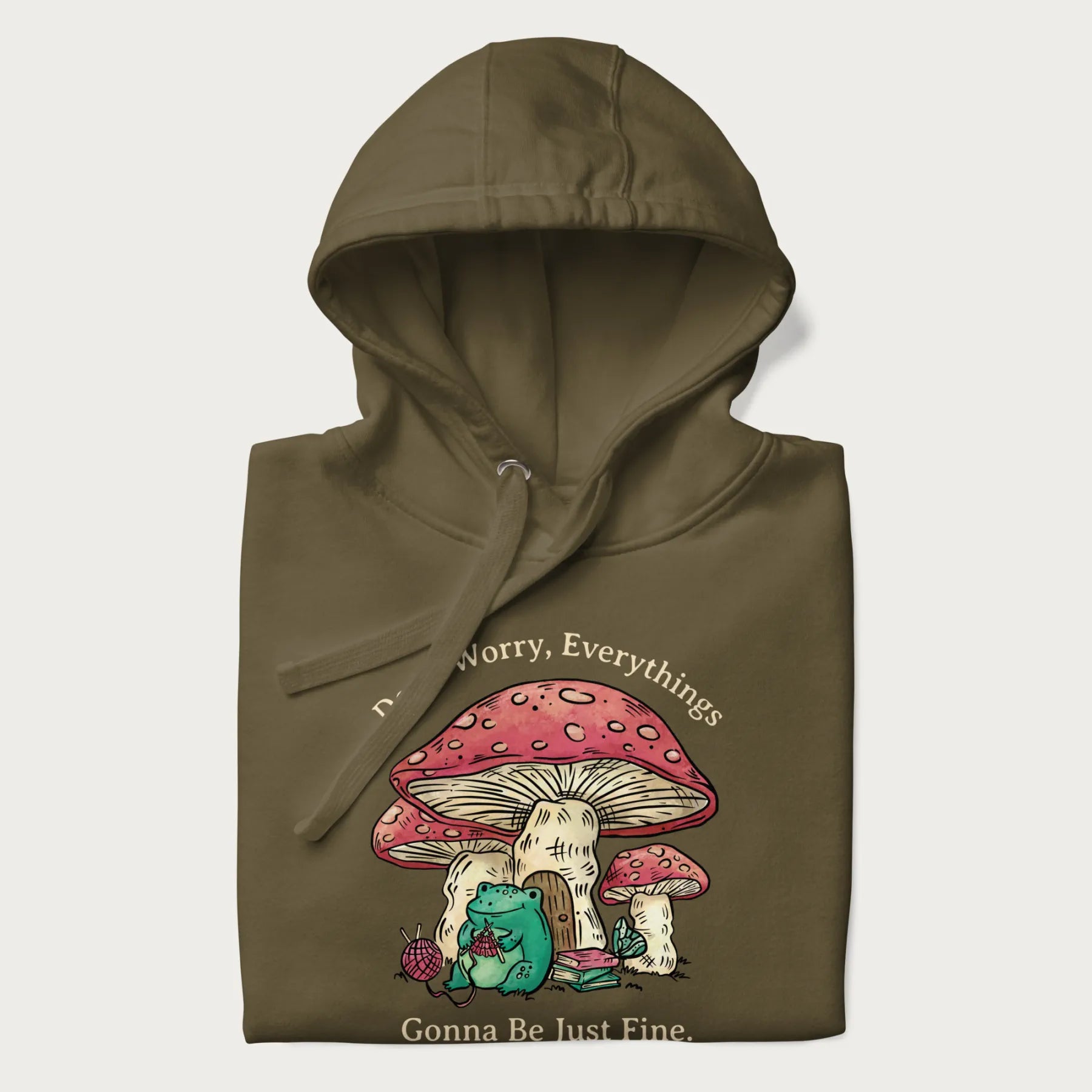 Folded military green hoodie with a graphic of a frog knitting under a large mushroom with the text 'Don't Worry, Everything's Gonna Be Just Fine'.