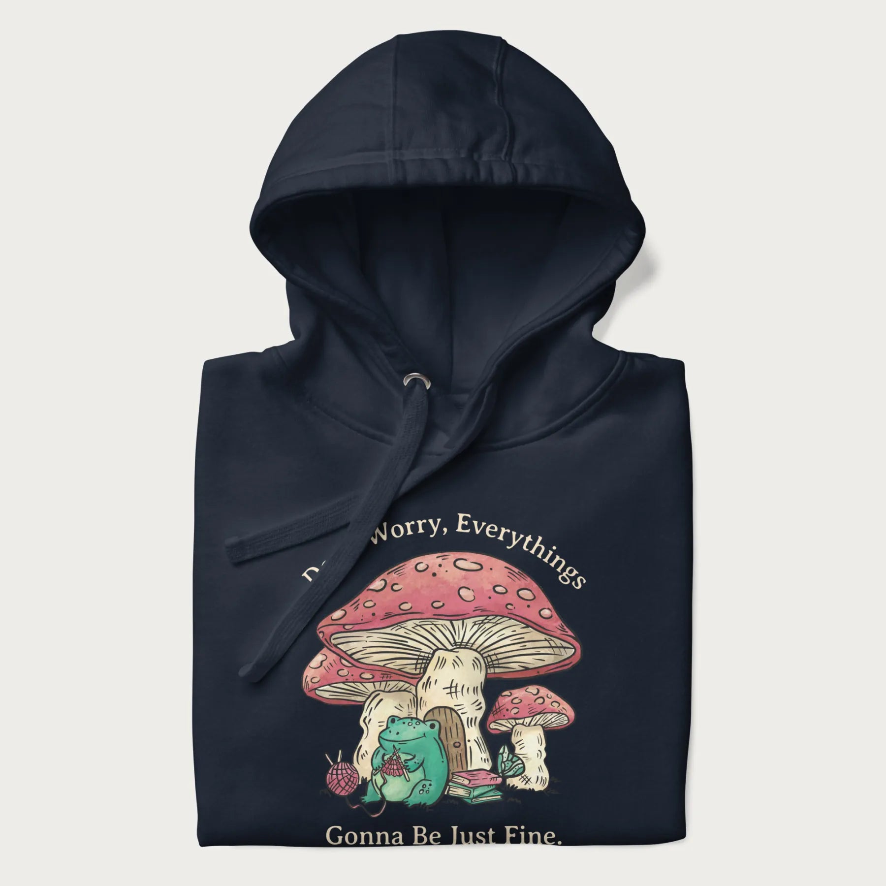 Folded navy blue hoodie with a graphic of a frog knitting under a large mushroom with the text 'Don't Worry, Everything's Gonna Be Just Fine'.