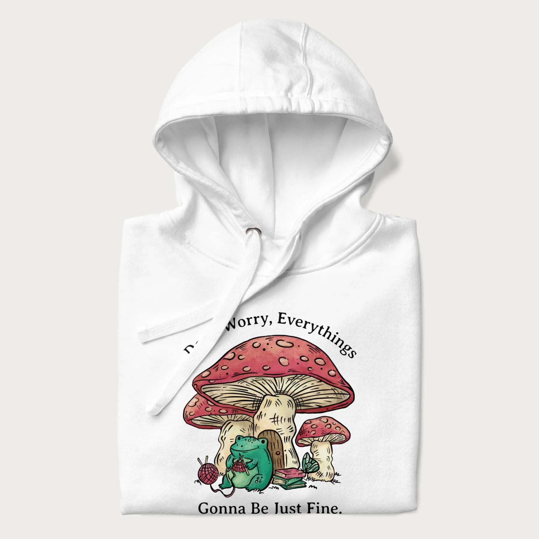 Folded white hoodie with a graphic of a frog knitting under a large mushroom with the text 'Don't Worry, Everything's Gonna Be Just Fine'.