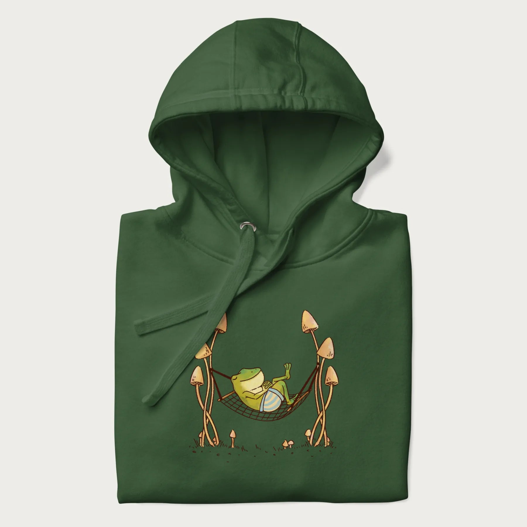 Folded forest green hoodie with a graphic of a frog lounging in a hammock between tall mushrooms.
