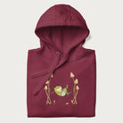 Folded maroon hoodie with a graphic of a frog lounging in a hammock between tall mushrooms.