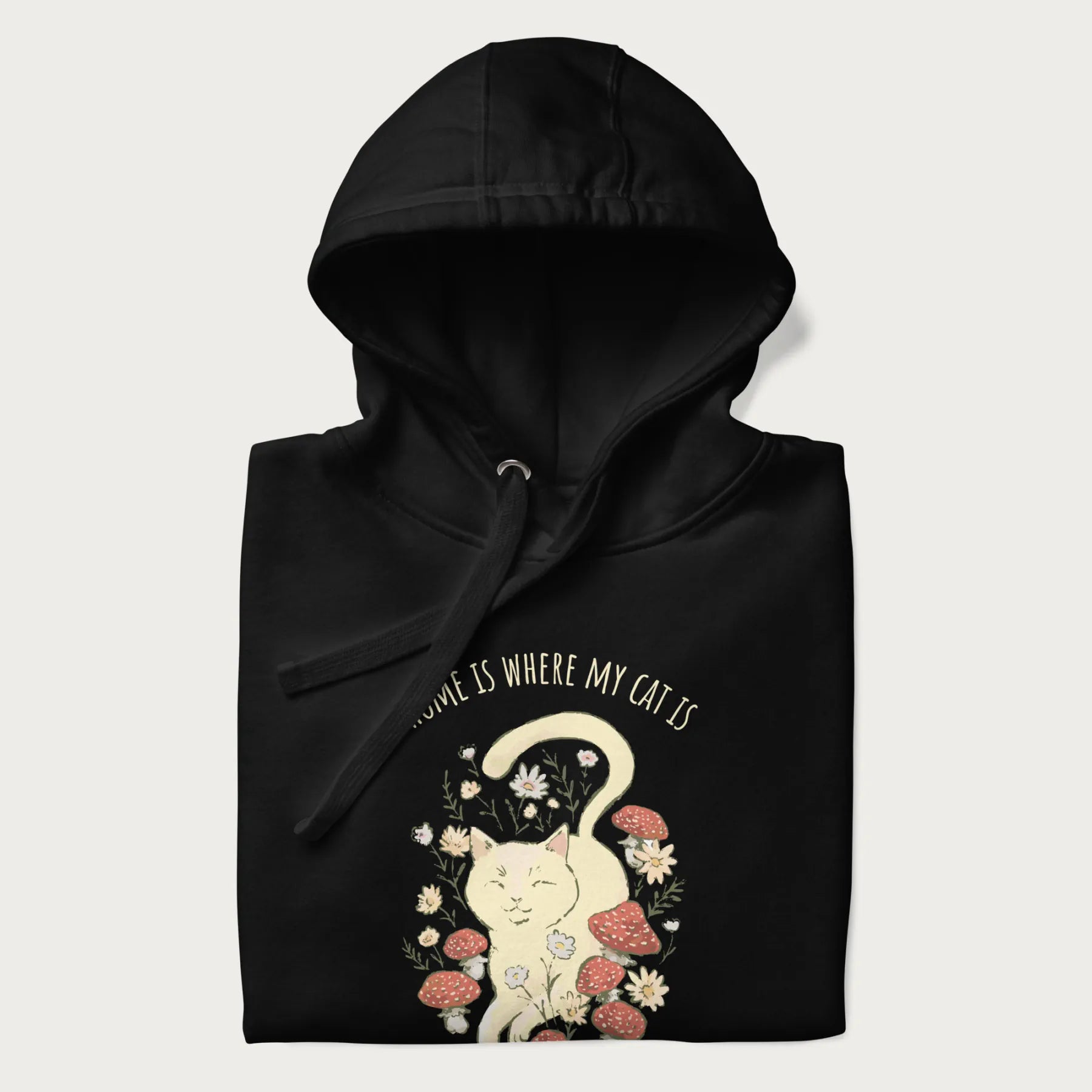 Folded black hoodie with a graphic of a cat nestled among flowers and mushrooms with the text 'Home is Where My Cat Is' above.