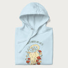 Folded light blue hoodie with a graphic of a cat nestled among flowers and mushrooms with the text 'Home is Where My Cat Is' above.