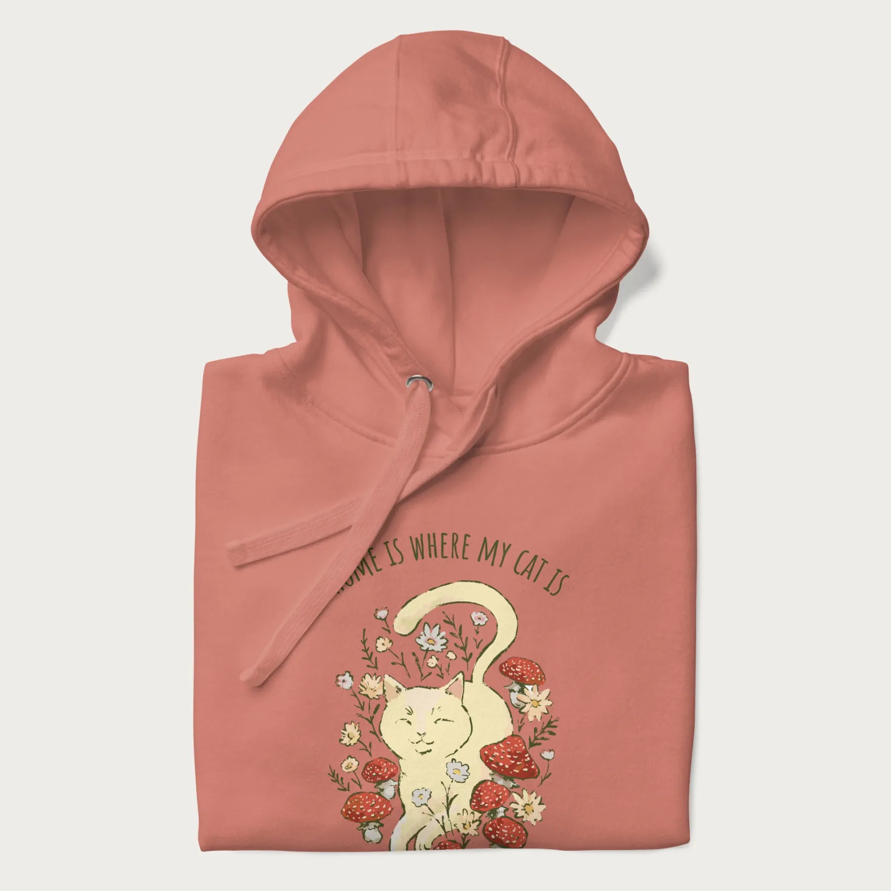 Folded light pink hoodie with a graphic of a cat nestled among flowers and mushrooms with the text 'Home is Where My Cat Is' above.