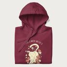 Folded maroon hoodie with a graphic of a cat nestled among flowers and mushrooms with the text 'Home is Where My Cat Is' above.