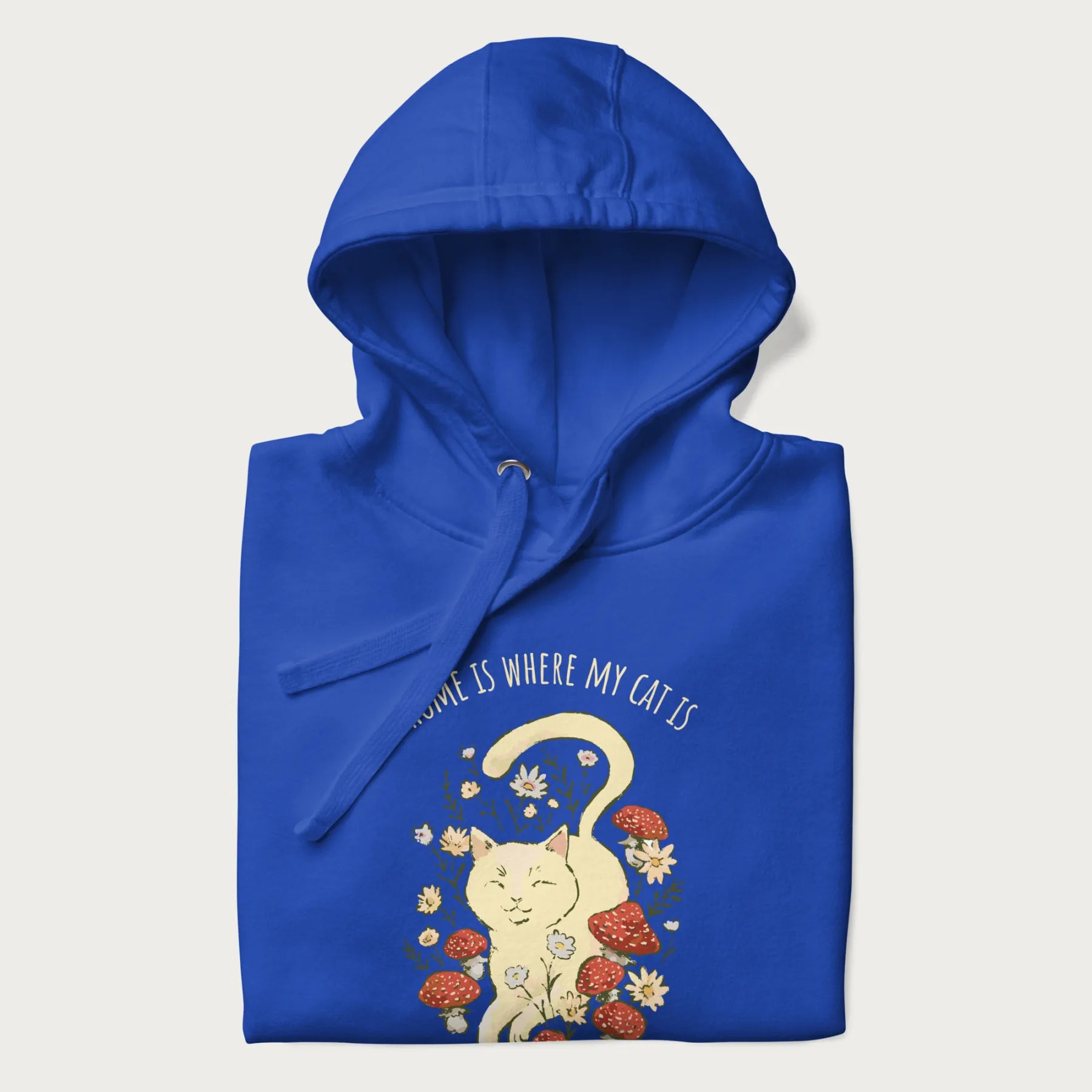 Folded royal blue hoodie with a graphic of a cat nestled among flowers and mushrooms with the text 'Home is Where My Cat Is' above.