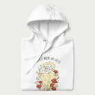 Folded white hoodie with a graphic of a cat nestled among flowers and mushrooms with the text 'Home is Where My Cat Is' above.