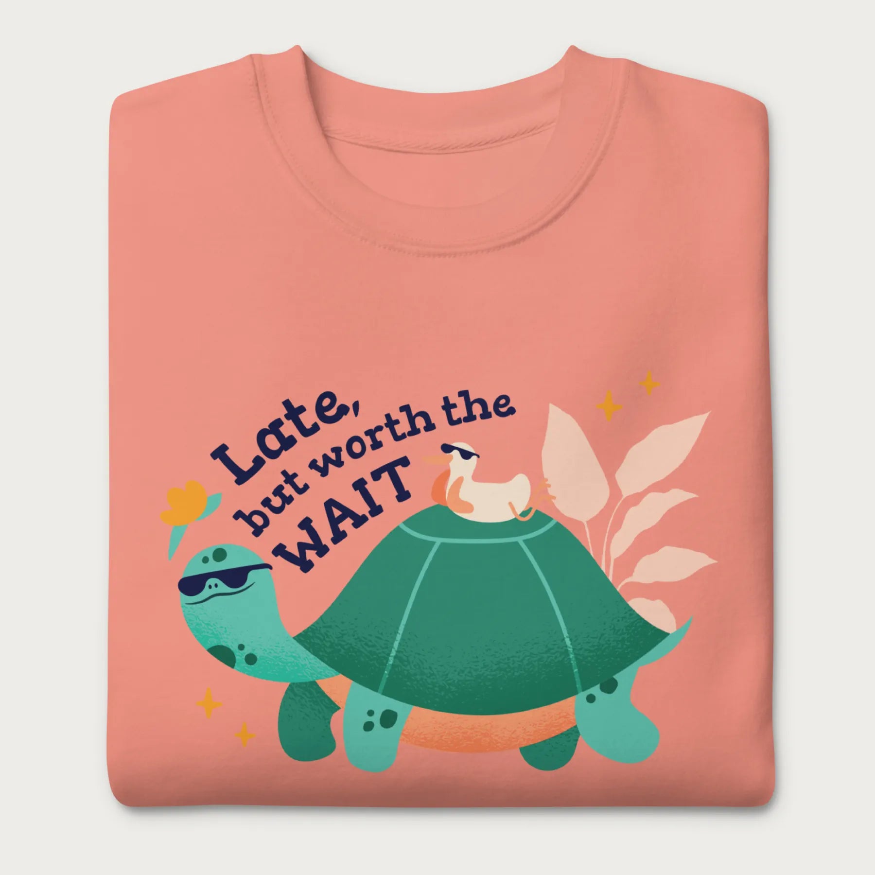 Folded light pink sweatshirt with a graphic of a turtle and bird wearing sunglasses with the phrase 'Late But Worth the Wait.'