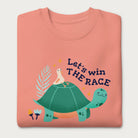 Folded light pink sweatshirt with a graphic of a turtle with a bird on it's shell with the phrase 'Let's Win the Race.'