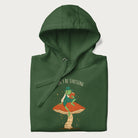 Folded forest green hoodie with graphic of a dapper frog reading a book on a mushroom with the text 'Don't Be Stressing, Life's a Blessing' above and below.