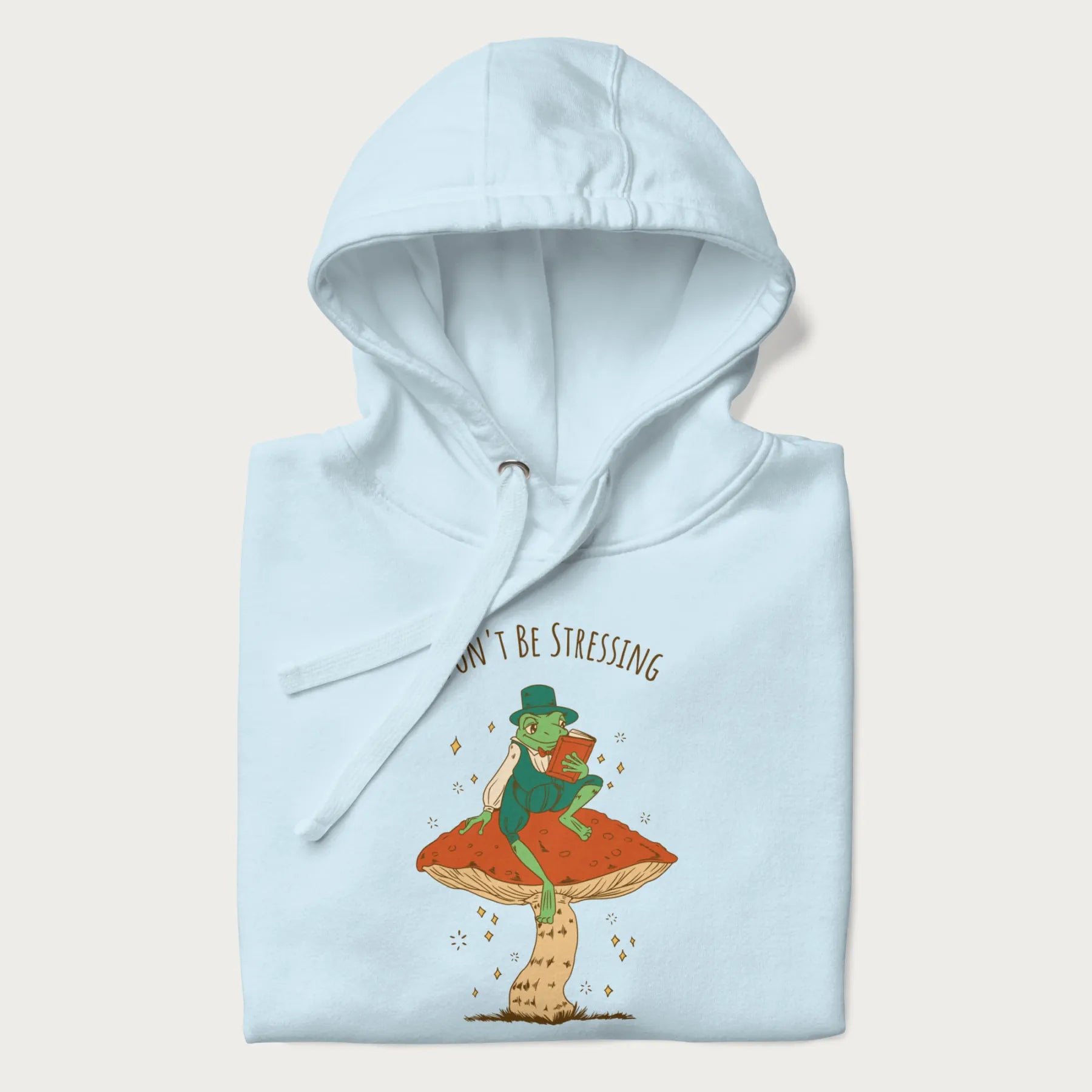 Folded light blue hoodie with graphic of a dapper frog reading a book on a mushroom with the text 'Don't Be Stressing, Life's a Blessing' above and below.