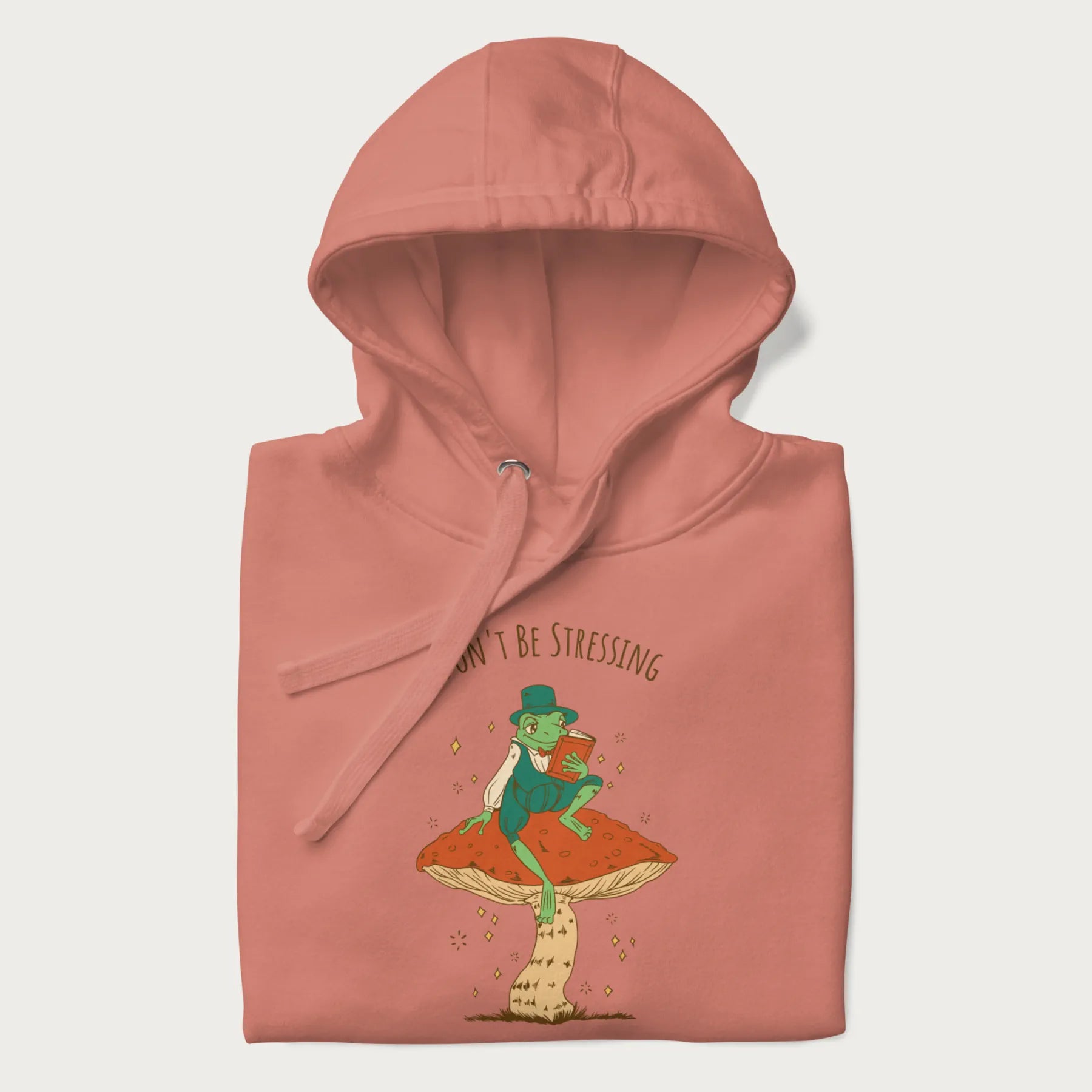 Folded light pink hoodie with graphic of a dapper frog reading a book on a mushroom with the text 'Don't Be Stressing, Life's a Blessing' above and below.