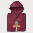 Folded maroon hoodie with graphic of a dapper frog reading a book on a mushroom with the text 'Don't Be Stressing, Life's a Blessing' above and below.