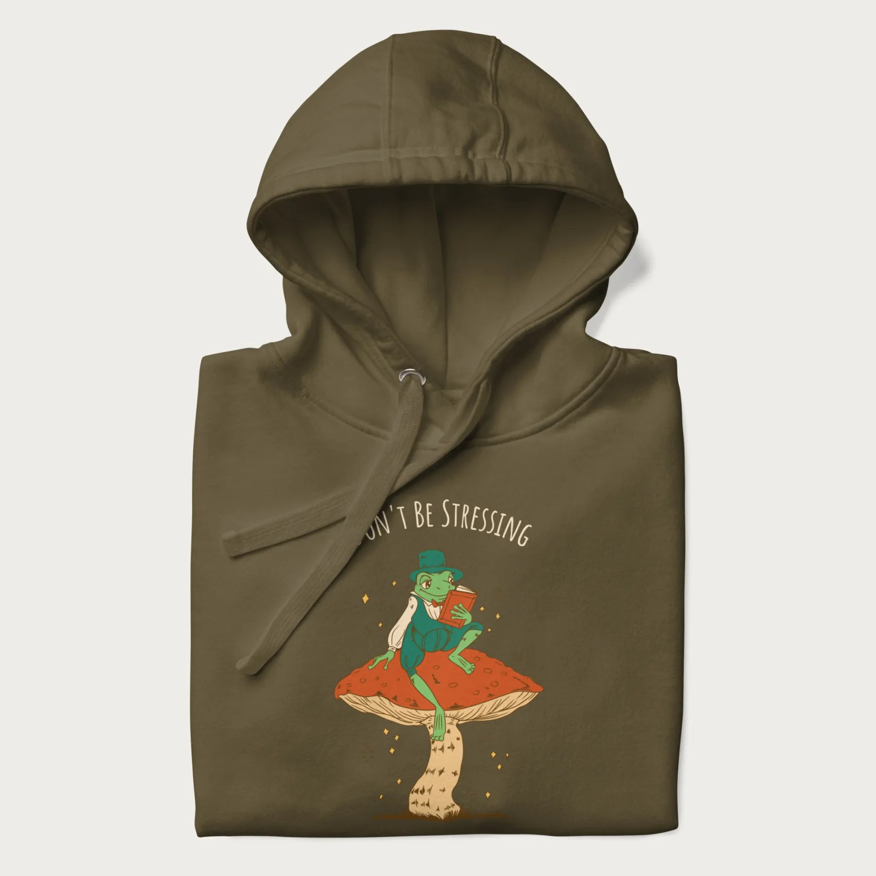 Folded military green hoodie with graphic of a dapper frog reading a book on a mushroom with the text 'Don't Be Stressing, Life's a Blessing' above and below.