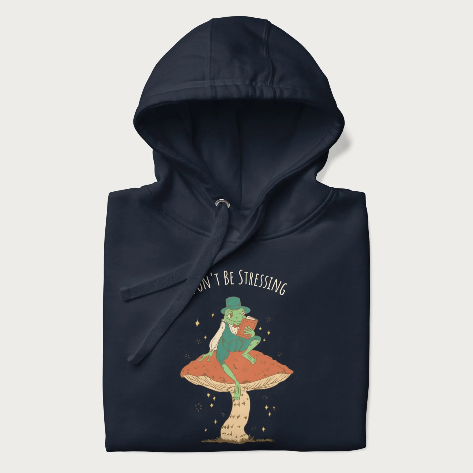 Folded navy blue hoodie with graphic of a dapper frog reading a book on a mushroom with the text 'Don't Be Stressing, Life's a Blessing' above and below.