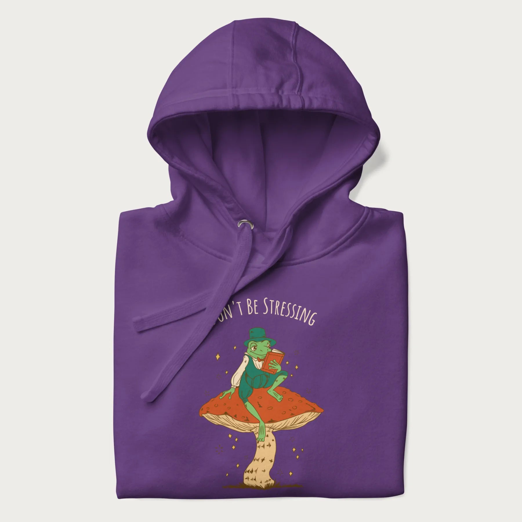 Folded purple hoodie with graphic of a dapper frog reading a book on a mushroom with the text 'Don't Be Stressing, Life's a Blessing' above and below.