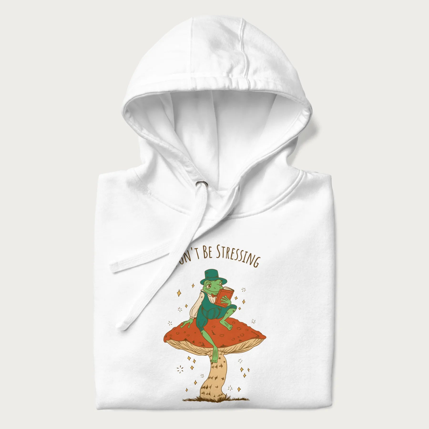 Folded white hoodie with graphic of a dapper frog reading a book on a mushroom with the text 'Don't Be Stressing, Life's a Blessing' above and below.