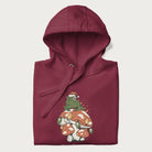 Folded maroon hoodie with graphic of a frog in a Santa hat on a mushroom with Christmas lights and the text 'Merry Christmas' below.