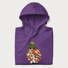 Folded purple hoodie with graphic of a frog in a Santa hat on a mushroom with Christmas lights and the text 'Merry Christmas' below.