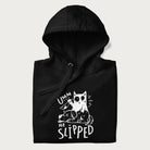 Folded black hoodie with a graphic of a white cat holding a knife over a dog with a funny phrase 'Umm, He Slipped'.