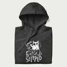Folded dark grey hoodie with a graphic of a white cat holding a knife over a dog with a funny phrase 'Umm, He Slipped'.