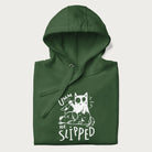 Folded green hoodie with a graphic of a white cat holding a knife over a dog with a funny phrase 'Umm, He Slipped'.