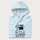 Folded light blue hoodie with a graphic of a black cat holding a knife over a dog with a funny phrase 'Umm, He Slipped'.