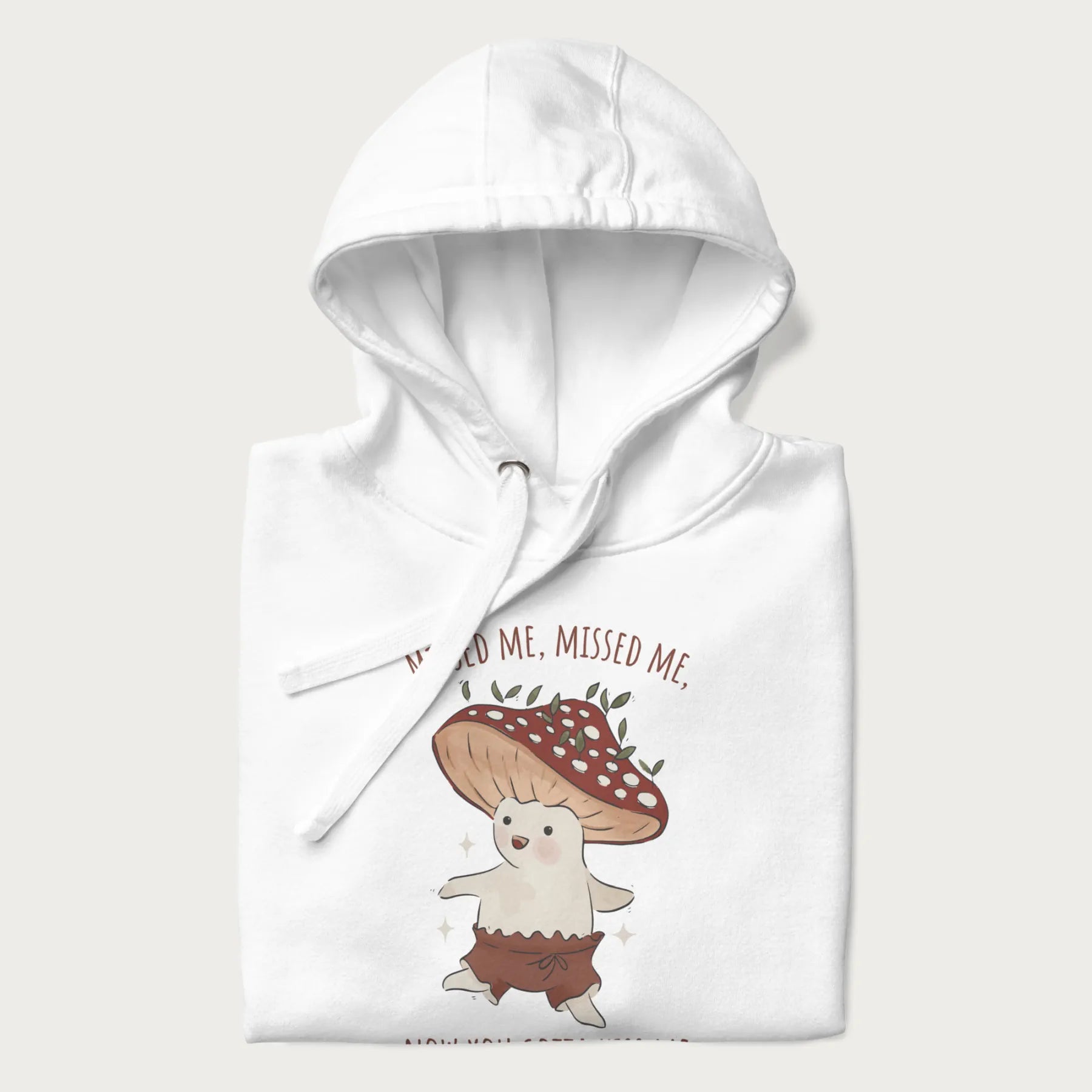 Folded white hoodie with a graphic of a cute cottagecore mushroom character dancing and the phrase 'Missed Me, Missed Me, Now You Gotta Kiss Me'.