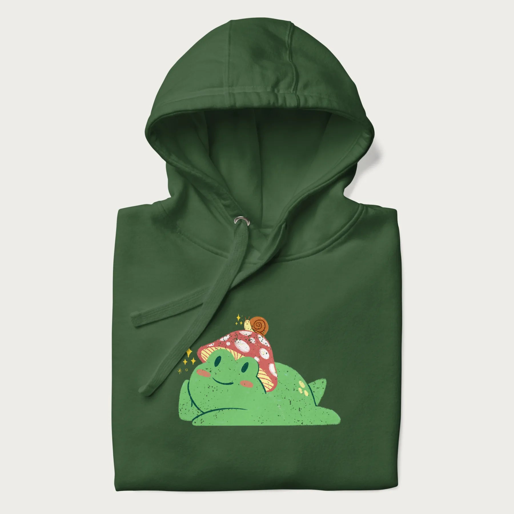 Folded forest green hoodie with a cottagecore design of a green frog wearing a mushroom cap and a snail on top.
