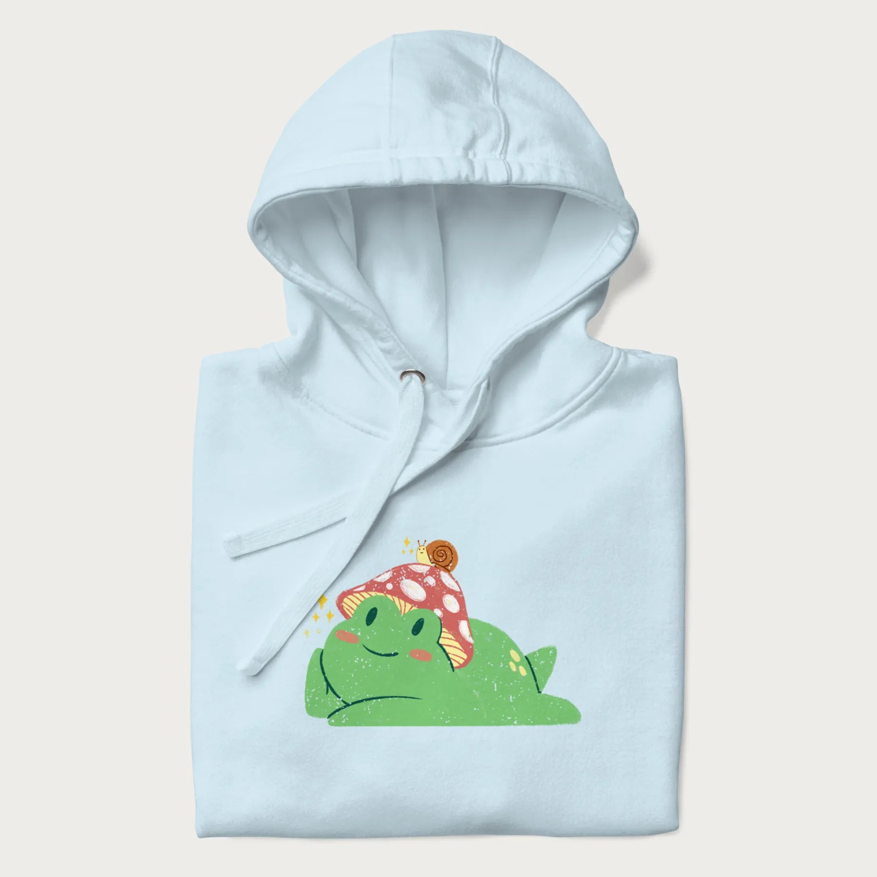 Folded light blue hoodie with a cottagecore design of a green frog wearing a mushroom cap and a snail on top.