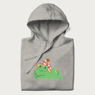 Folded light grey hoodie with a cottagecore design of a green frog wearing a mushroom cap and a snail on top.