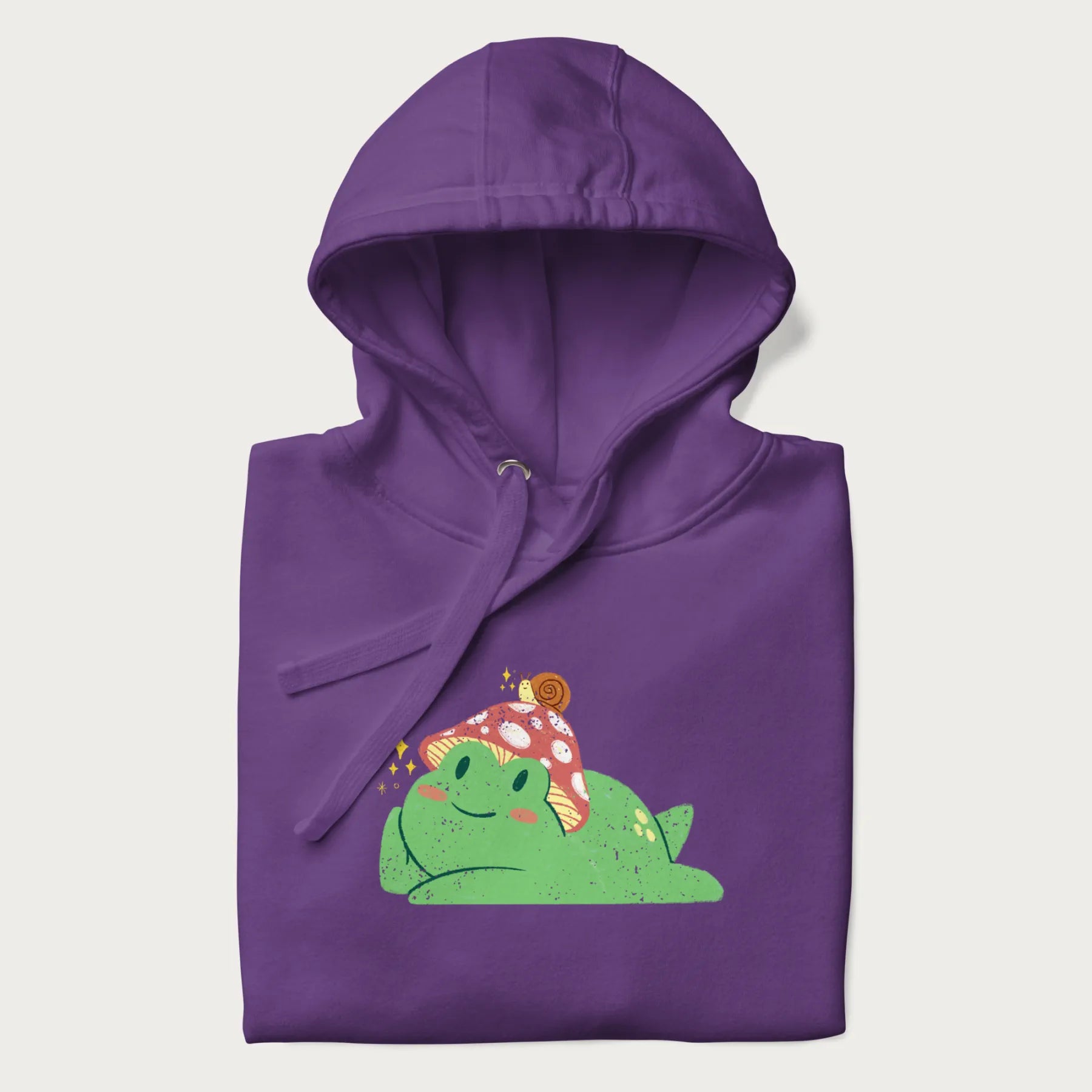 Folded purple hoodie with a cottagecore design of a green frog wearing a mushroom cap and a snail on top.