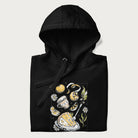 Folded black hoodie featuring a Cottagecore-themed graphic with oranges, a floral porcelain cup, orange pie, and daisies.