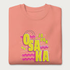 Folded light pink sweatshirt with a vibrant graphic of the words "OSAKA JAPAN" in large yellow letters, surrounded by pink zigzag lines.