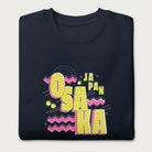 Folded navy blue sweatshirt with a vibrant graphic of the words "OSAKA JAPAN" in large yellow letters, surrounded by pink zigzag lines.