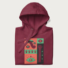 Folded maroon hoodie with mushroom psychedelic designs of surreal elements like lips, eyes, and mushrooms.