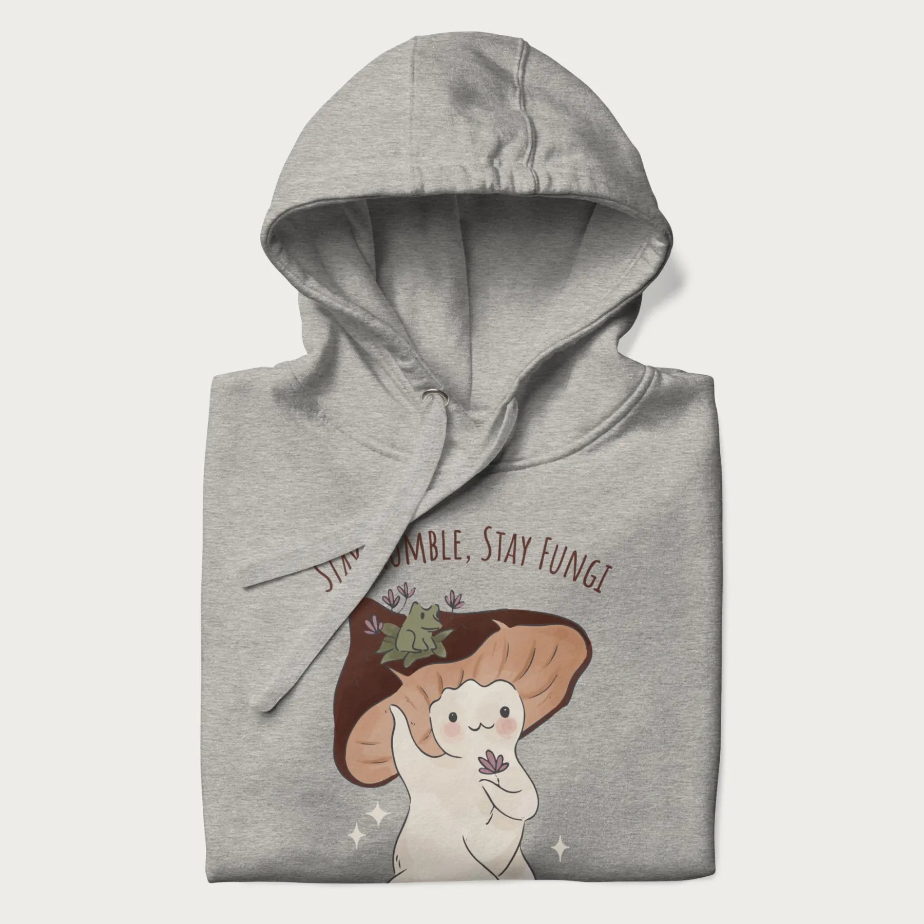 Folded light grey hoodie with graphic of a cute mushroom character holding a flower with a frog on its cap and the text 'Stay Humble, Stay Fungi' above.