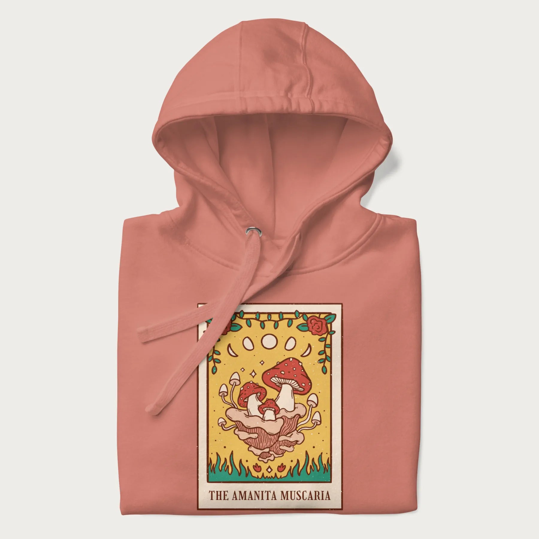 Folded light pink hoodie featuring a graphic of Amanita Muscaria mushrooms with floral accents and moon phases, resembling a tarot card.