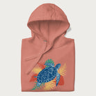 Folded light pink hoodie with a vibrant graphic of a blue sea turtle and colorful tropical leaves.