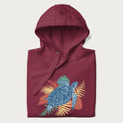 Folded maroon hoodie with a vibrant graphic of a blue sea turtle and colorful tropical leaves.