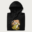 Folded black hoodie with cottagecore graphic of a frog under mushrooms with foliage in the background.