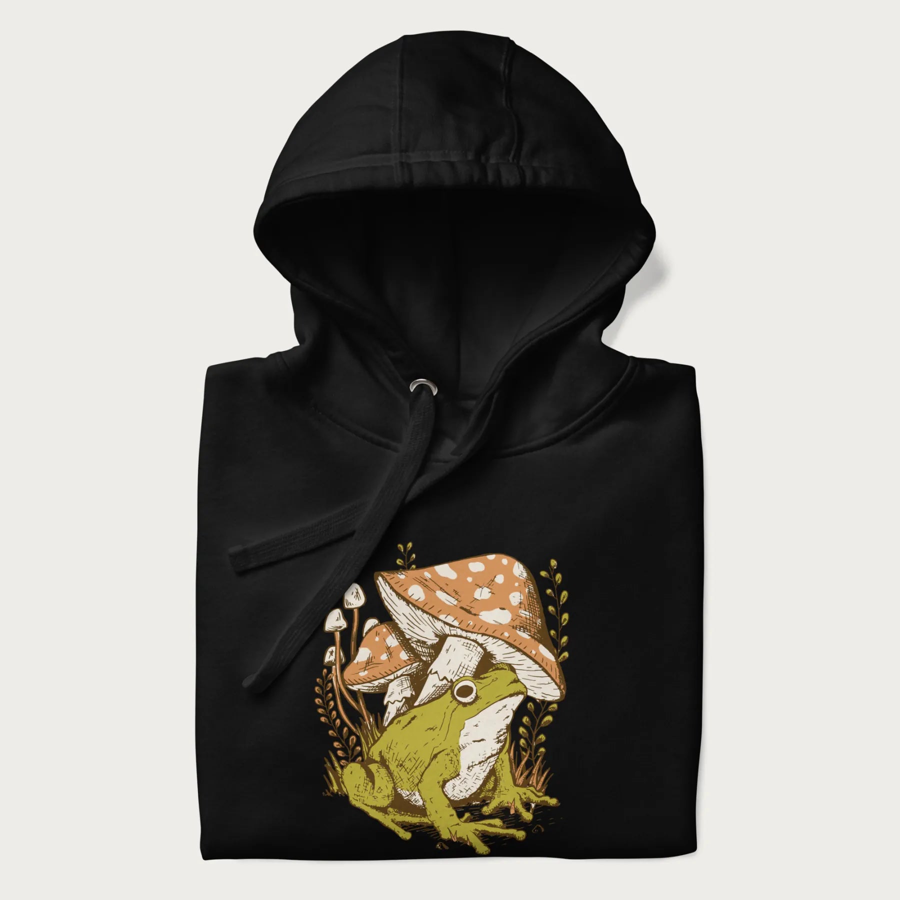 Folded black hoodie with cottagecore graphic of a frog under mushrooms with foliage in the background.