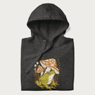 Folded dark grey hoodie with cottagecore graphic of a frog under mushrooms with foliage in the background.