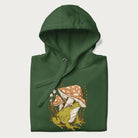 Folded forest green hoodie with cottagecore graphic of a frog under mushrooms with foliage in the background.