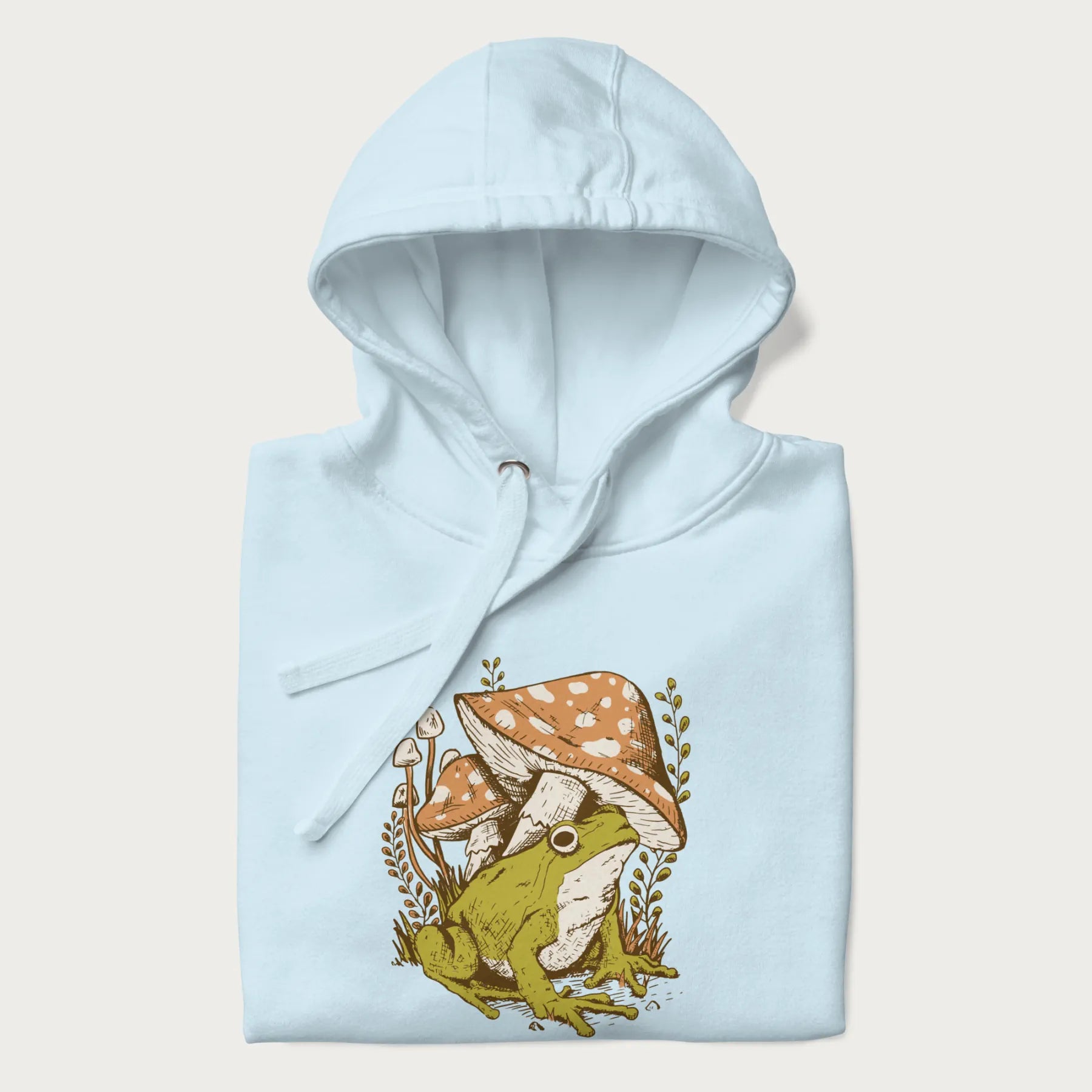 Folded light blue hoodie with cottagecore graphic of a frog under mushrooms with foliage in the background.