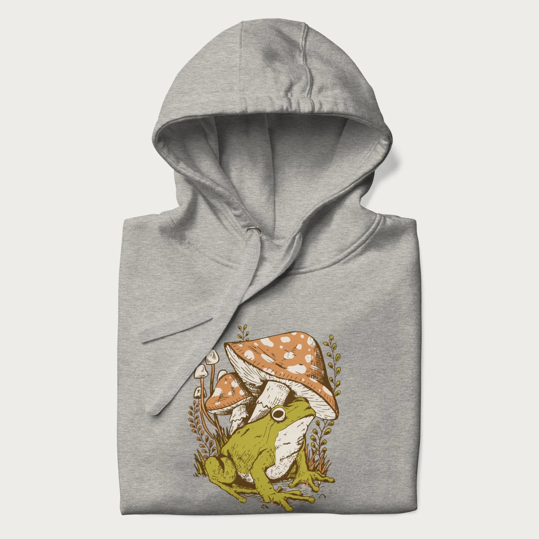 Folded light grey hoodie with cottagecore graphic of a frog under mushrooms with foliage in the background.