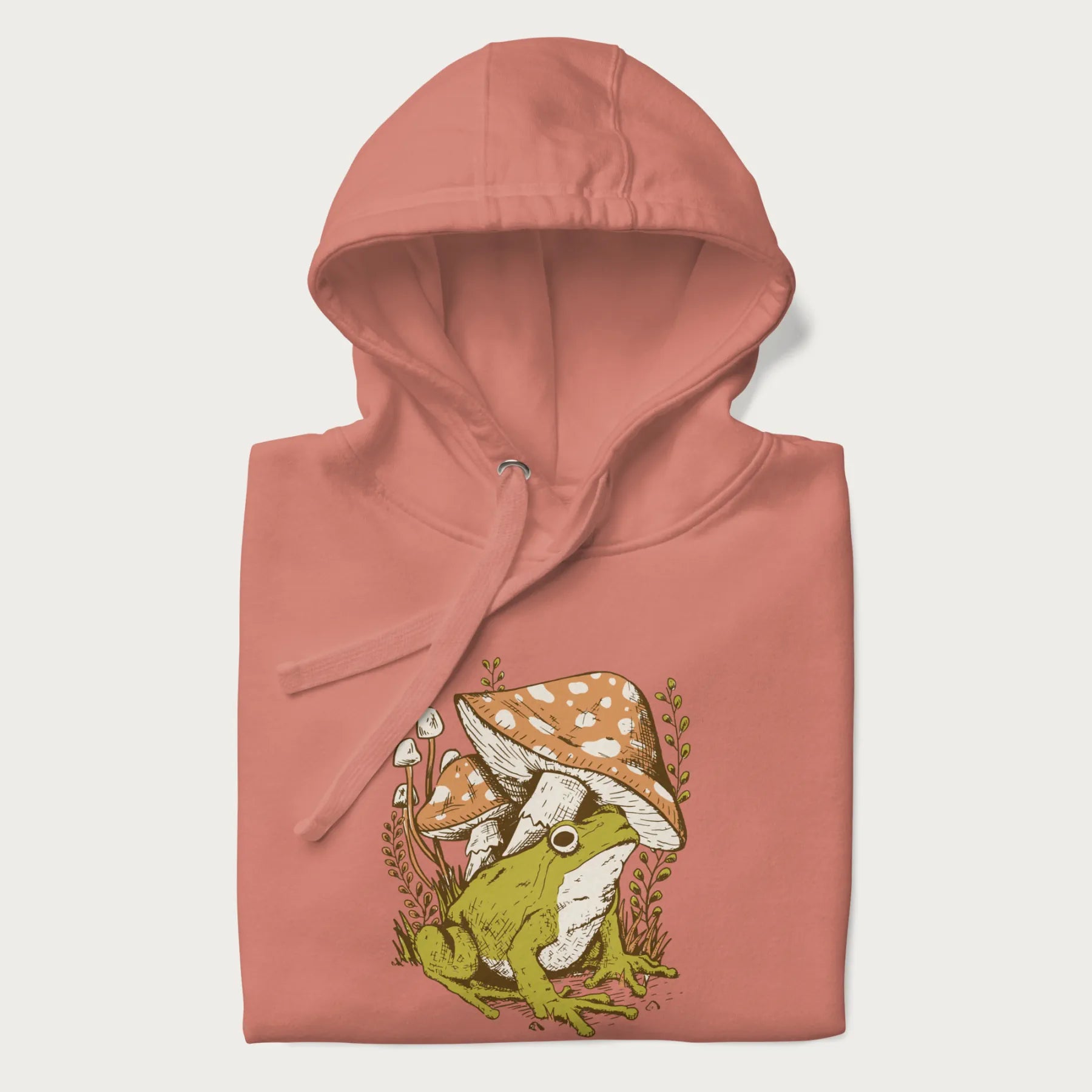 Folded light pink hoodie with cottagecore graphic of a frog under mushrooms with foliage in the background.